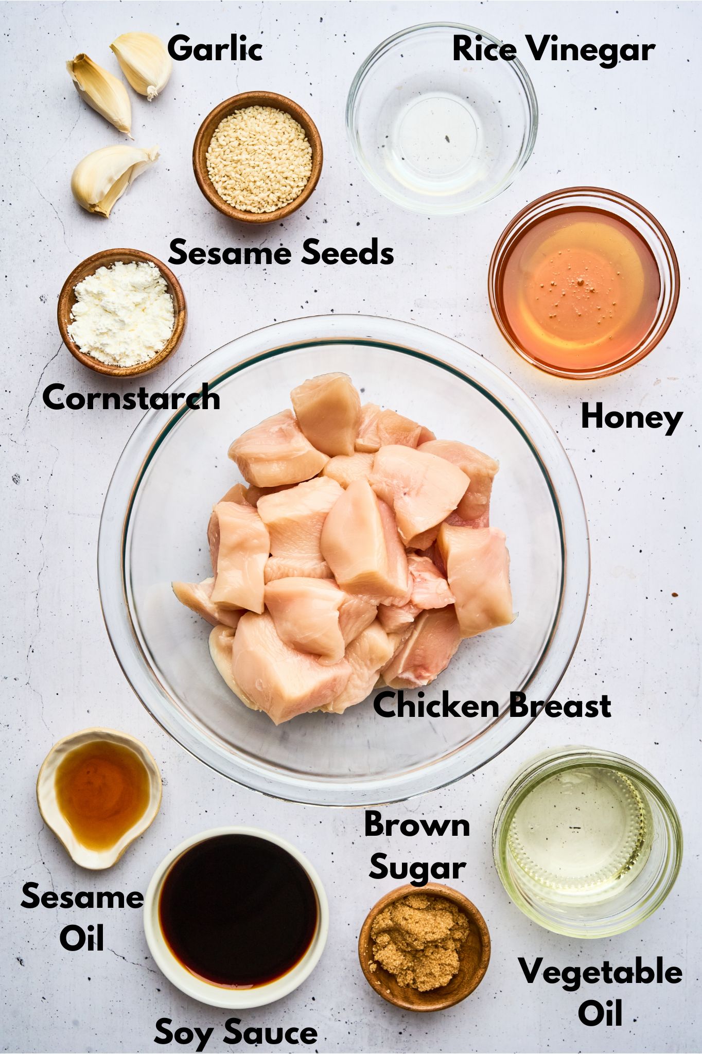 Overhead view of the ingredients for honey sesame chicken, including raw chicken chunks, garlic, sesame seeds, cornstarch, soy sauce, honey, sesame oil, rice vinegar, and brown sugar, arranged around a bowl.