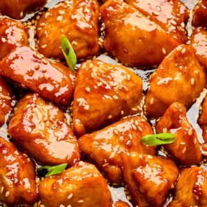 Close-up of honey sesame chicken pieces coated in a glossy, golden sauce with sesame seeds and green onion garnish.