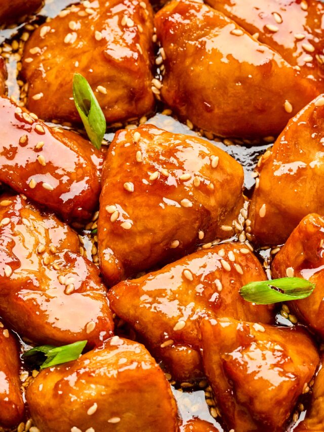 Close-up of honey sesame chicken pieces coated in a glossy, golden sauce with sesame seeds and green onion garnish.