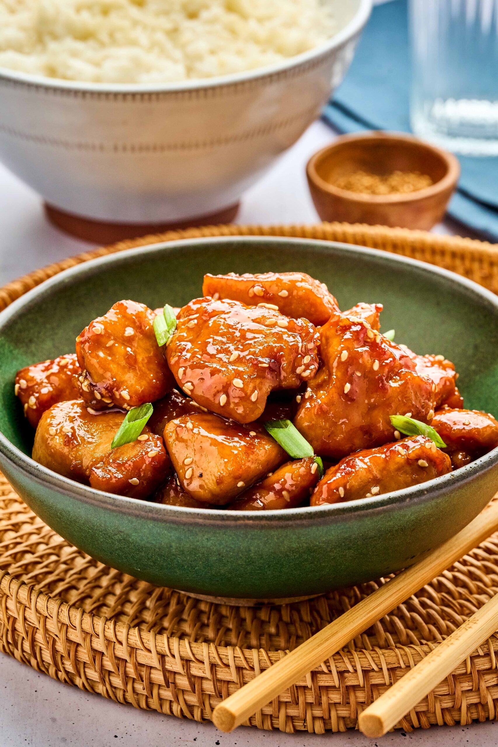 Bowl of honey sesame chicken garnished with green onions, served with rice in the background and chopsticks placed beside the bowl.