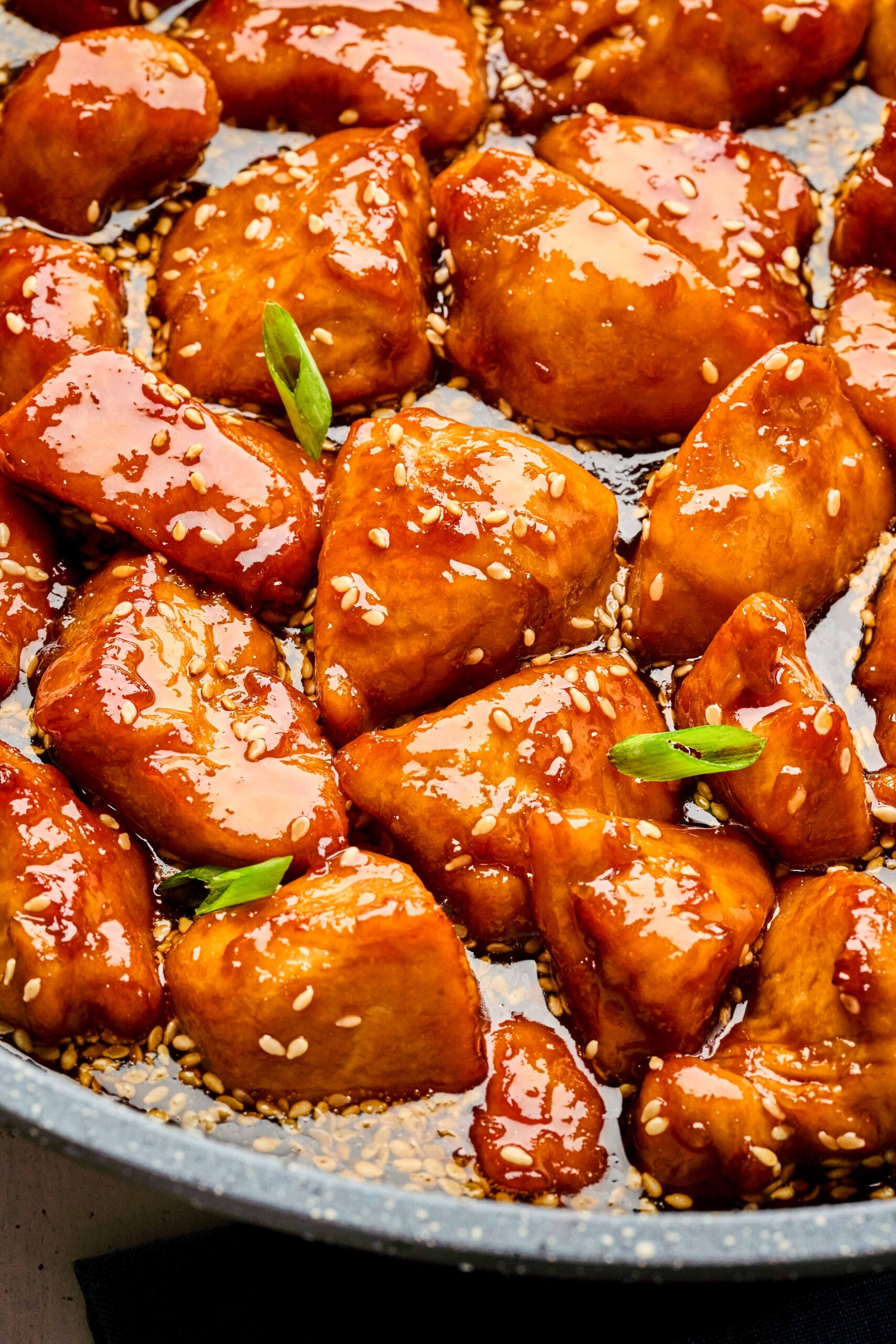 Close-up of honey sesame chicken pieces coated in a glossy, golden sauce with sesame seeds and green onion garnish.