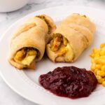 Two crescent rolls filled with Thanksgiving leftovers, including turkey, stuffing, and mashed potatoes, served on a plate with cranberry sauce and corn.