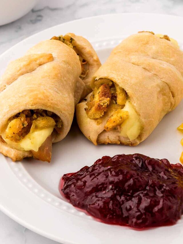Two crescent rolls filled with Thanksgiving leftovers, including turkey, stuffing, and mashed potatoes, served on a plate with cranberry sauce and corn.