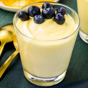 A glass of creamy lemon mousse topped with fresh blueberries, with a plate of candied lemon slices in the background, on a dark green surface accompanied by golden spoons.