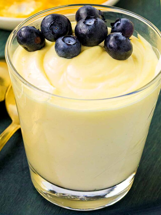 A glass of creamy lemon mousse topped with fresh blueberries, with a plate of candied lemon slices in the background, on a dark green surface accompanied by golden spoons.
