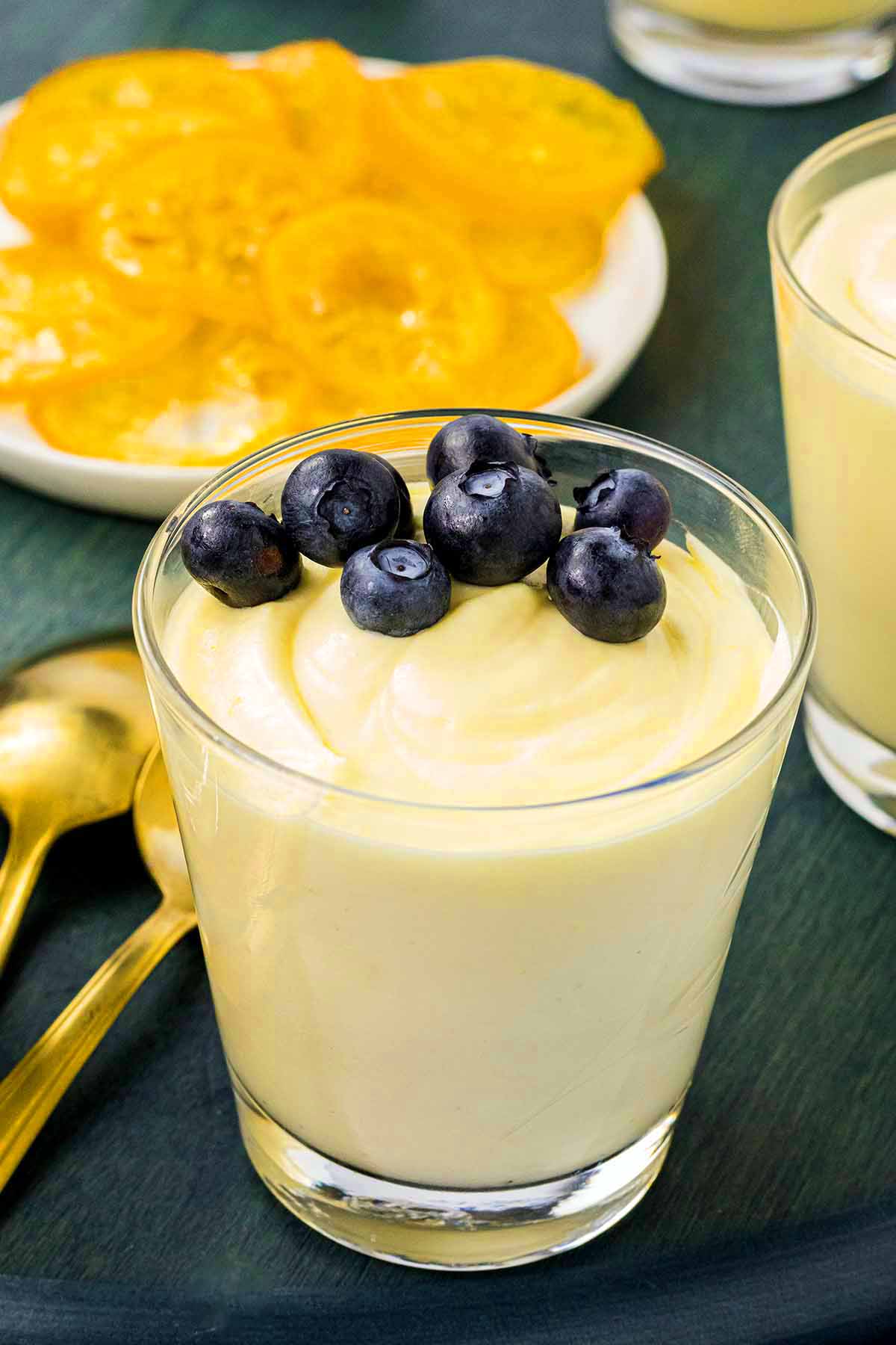 A glass of smooth lemon mousse topped with fresh blueberries, with a plate of candied lemon slices in the background and golden spoons on the side.