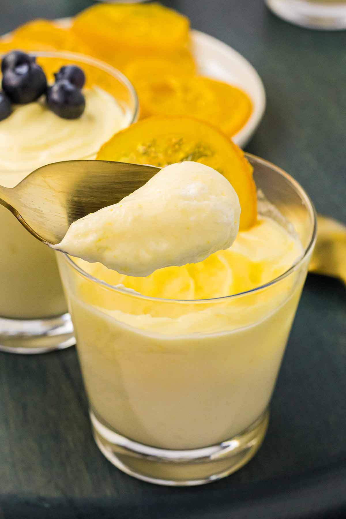 A spoonful of light and fluffy lemon mousse being lifted from a glass, garnished with a slice of candied lemon, with another glass of mousse topped with blueberries in the background.