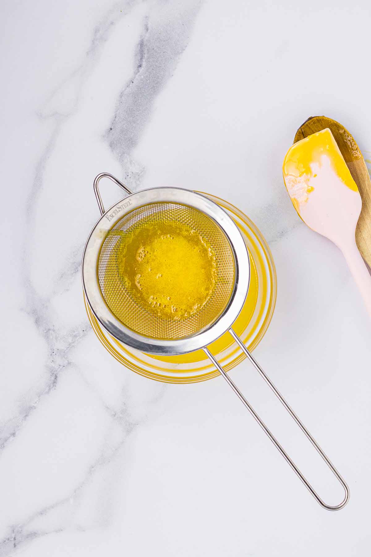 Lemon curd being strained into a bowl with a spatula and wooden spoon beside it. Step 5 of making lemon curd for lemon mousse.