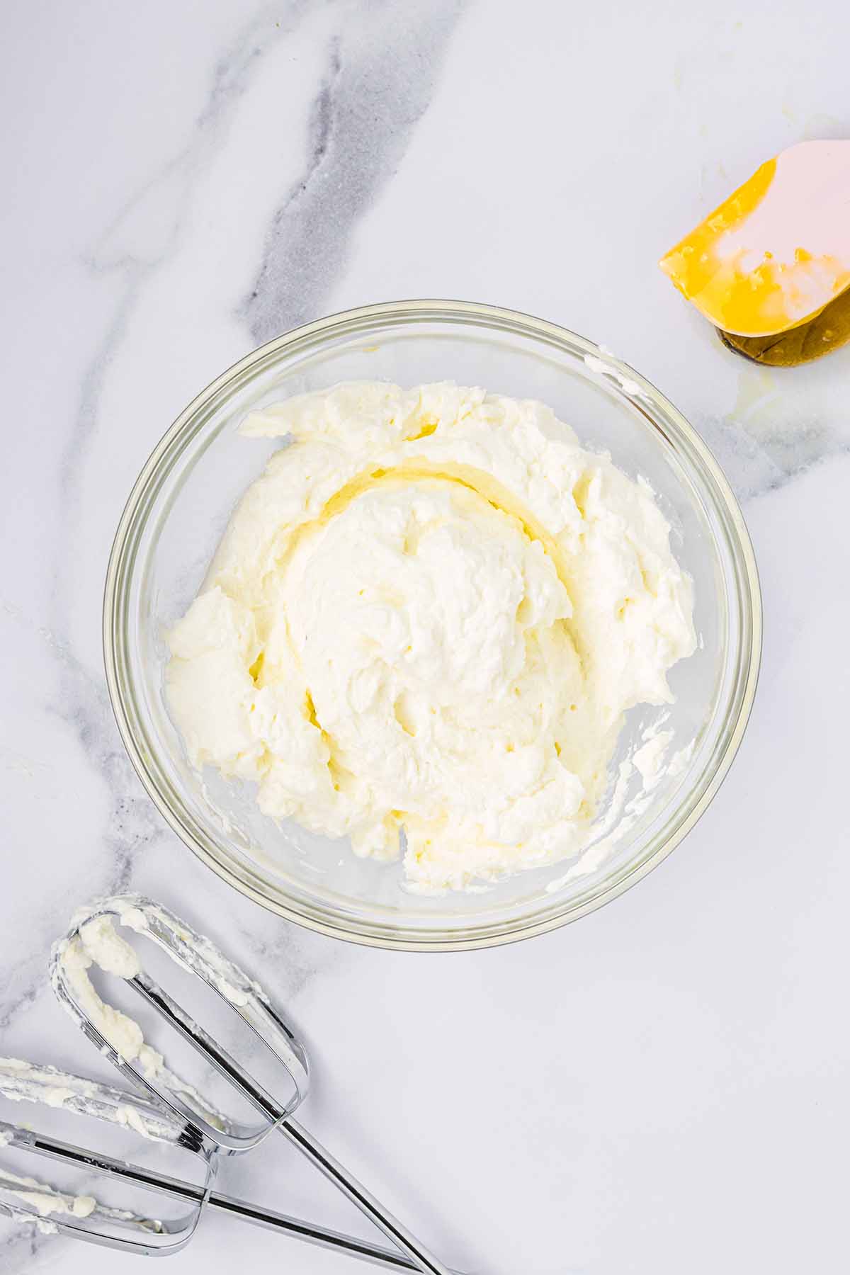 Whipped cream in a bowl with beaters and a spatula beside it. Step 6 of making lemon mousse.