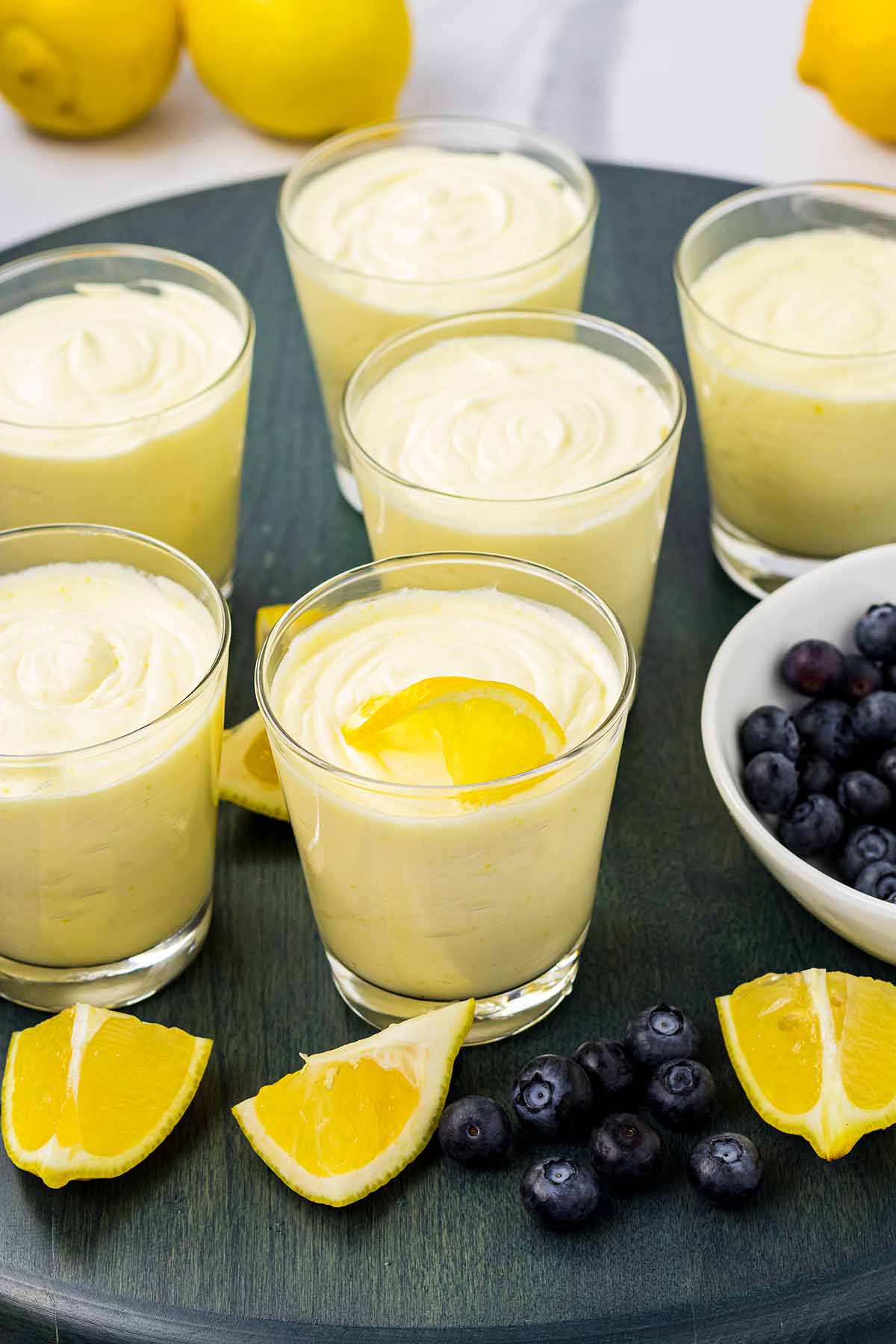 Six glasses of lemon mousse, one garnished with a lemon slice, arranged on a dark tray with fresh lemon wedges and blueberries nearby. Final step of the lemon mousse recipe.