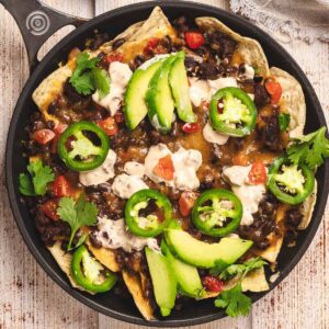 Loaded beef nachos topped with sliced avocado, jalapeños, tomatoes, sour cream, and fresh cilantro served in a black skillet.