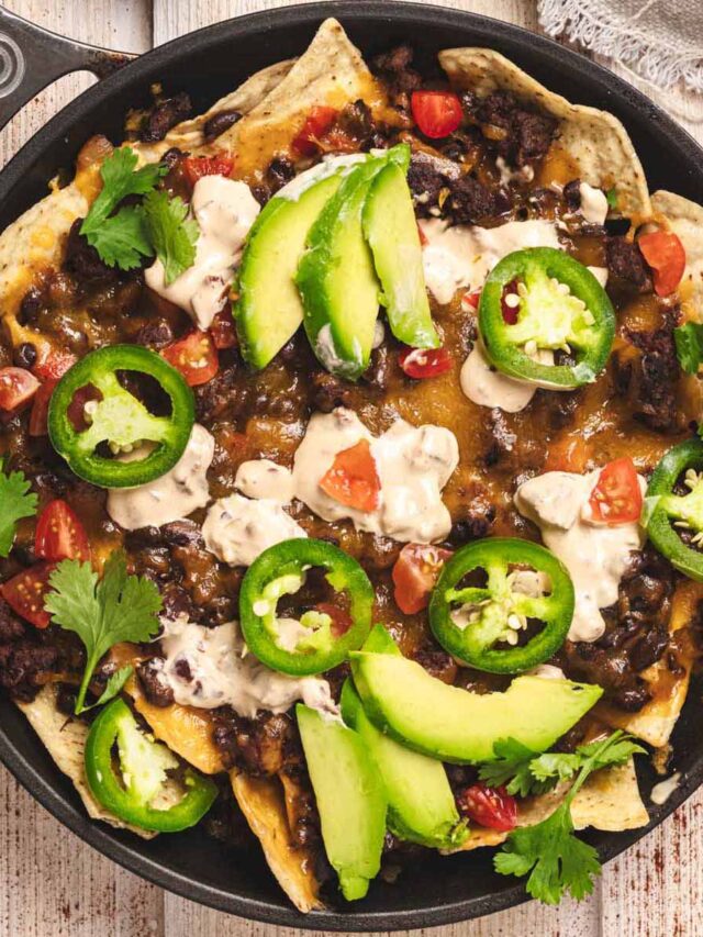 Loaded beef nachos topped with sliced avocado, jalapeños, tomatoes, sour cream, and fresh cilantro served in a black skillet.