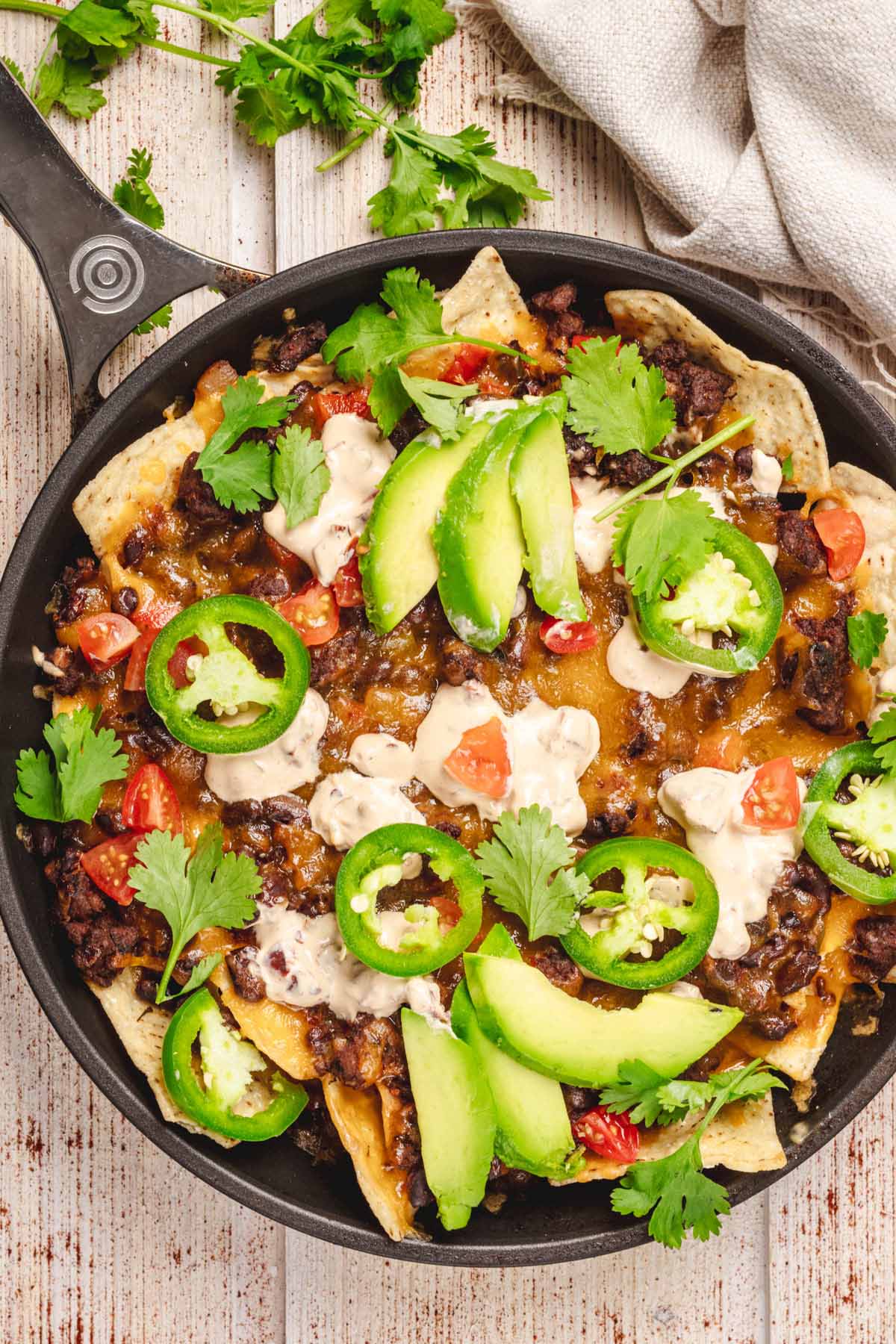 Close-up of loaded beef nachos topped with melted cheese, sliced avocado, jalapeños, fresh cilantro, diced tomatoes, and sour cream, served in a black skillet.