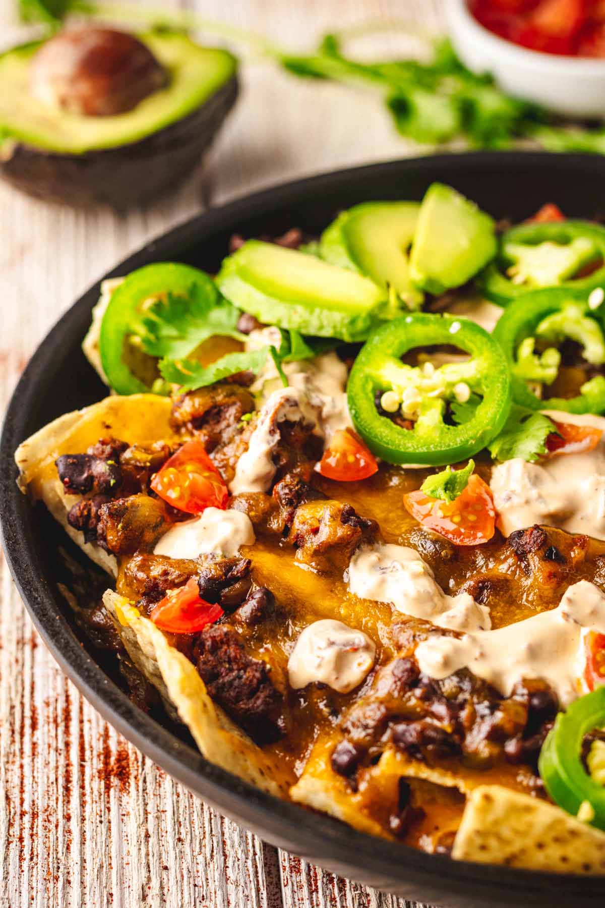 Close-up of loaded beef nachos with melted cheese, ground beef, diced tomatoes, jalapeños, sour cream, and sliced avocado in a black skillet, with an avocado and fresh cilantro in the background.