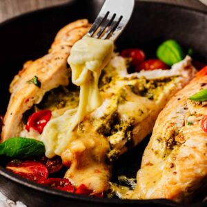 Close-up of a mozzarella-stuffed chicken breast cut in half, with melted cheese and pesto oozing out, served alongside roasted cherry tomatoes with melted cheese being pulled away by a fork in a black skillet.