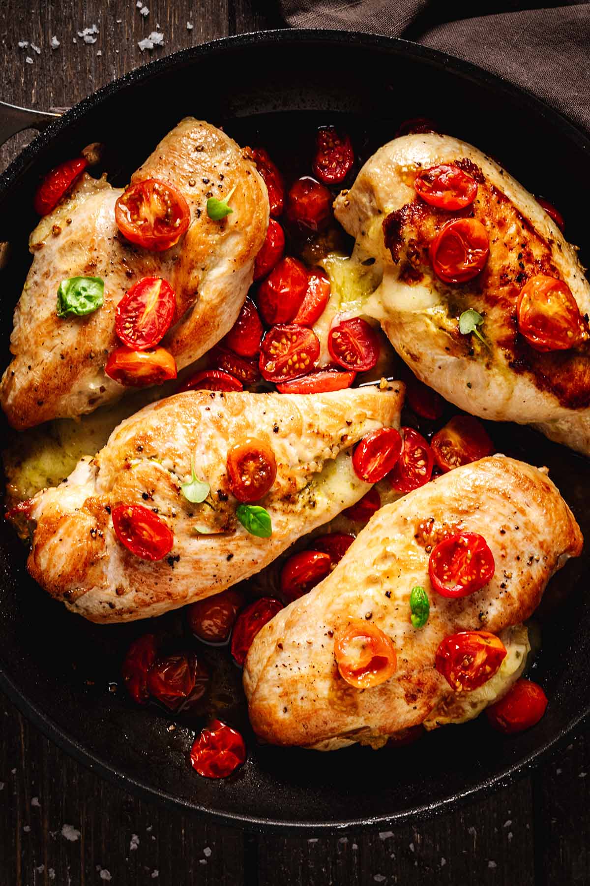 Top view of four mozzarella-stuffed chicken breasts baked with roasted cherry tomatoes and garnished with fresh basil in a black skillet, with melted cheese visible from inside the chicken.