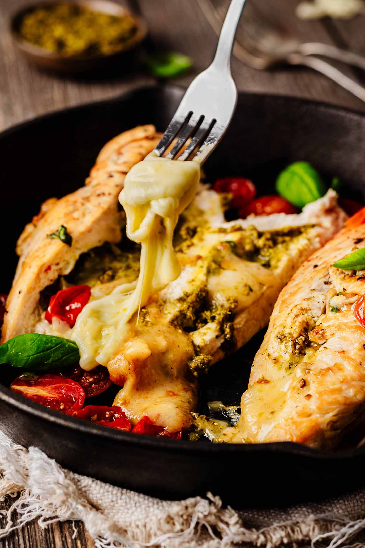 Close-up of a mozzarella-stuffed chicken breast cut in half, with melted cheese and pesto oozing out, served alongside roasted cherry tomatoes with melted cheese being pulled away by a fork in a black skillet.