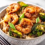 Close-up of shrimp and broccoli stir fry served over rice in a white bowl, garnished with sesame seeds and green onions.