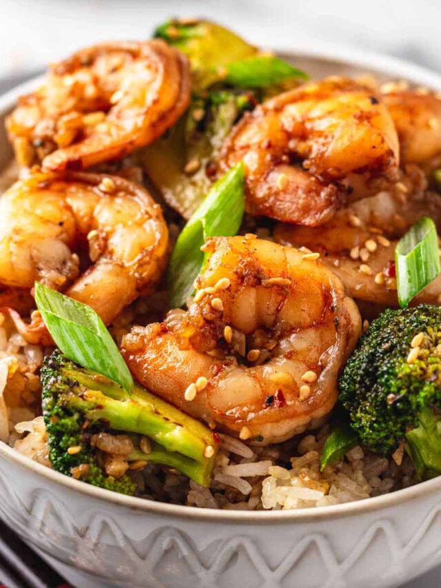 Close-up of shrimp and broccoli stir fry served over rice in a white bowl, garnished with sesame seeds and green onions.