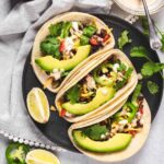 Soft beef tacos topped with sliced avocado, jalapeños, cilantro, and cheese on a dark plate, with lime wedges on the side and a light cloth napkin in the background.