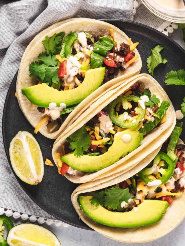 Soft beef tacos topped with sliced avocado, jalape&ntilde;os, cilantro, and cheese on a dark plate, with lime wedges on the side and a light cloth napkin in the background.
