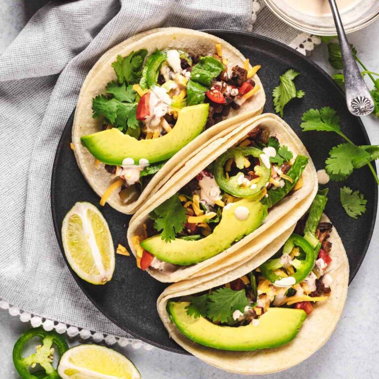 Soft beef tacos topped with sliced avocado, jalapeños, cilantro, and cheese on a dark plate, with lime wedges on the side and a light cloth napkin in the background.