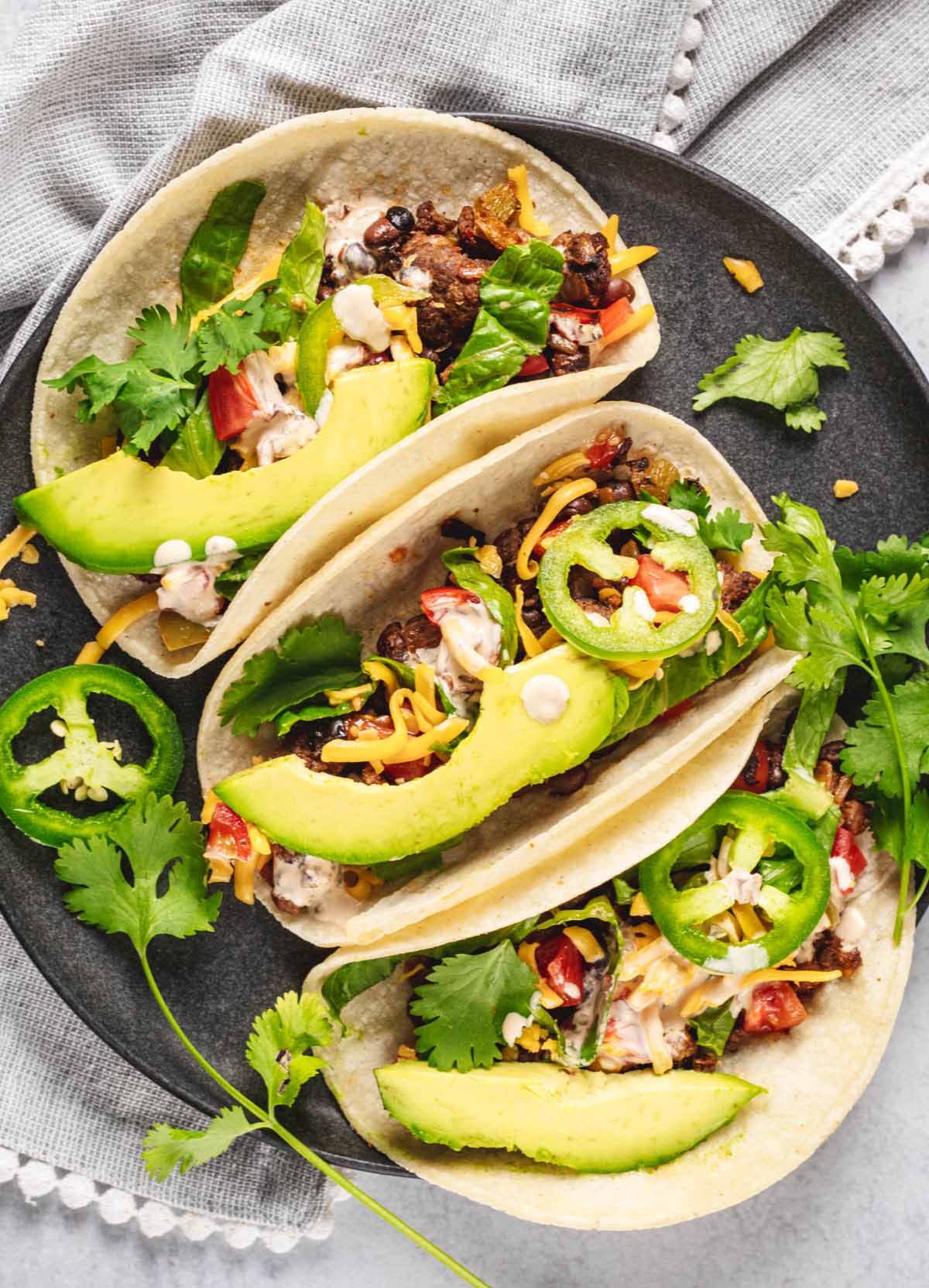 Soft beef tacos topped with sliced avocado, jalapeños, cheese, and cilantro on a black plate, with a light gray napkin and fresh cilantro in the background.