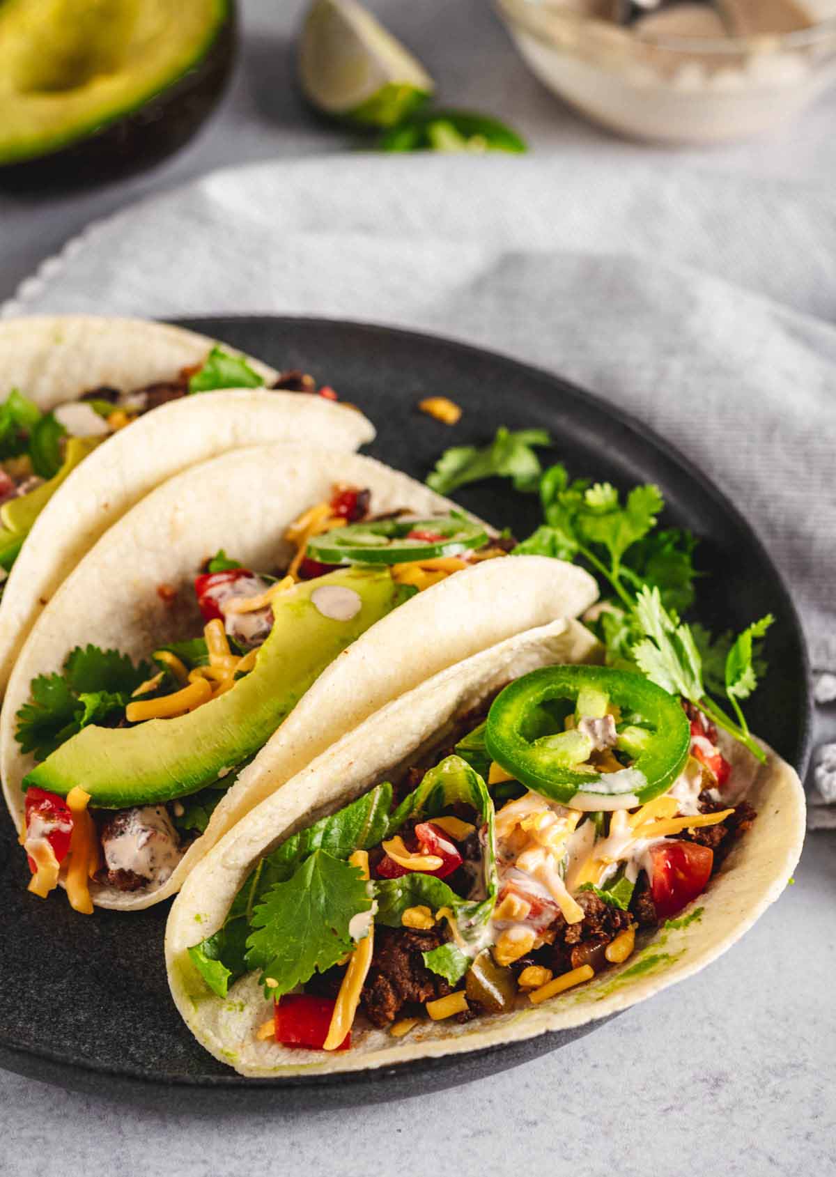 Soft beef tacos filled with seasoned beef, avocado, jalapeños, cheese, and cilantro on a black plate, with lime and sauce in the background.