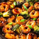 A close-up of teriyaki shrimp stir fry with plump shrimp and broccoli coated in glossy sauce, garnished with sesame seeds and green onions in a dark skillet.