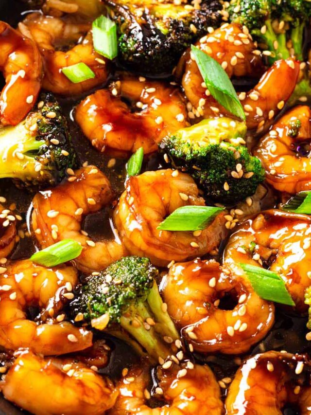 A close-up of teriyaki shrimp stir fry with plump shrimp and broccoli coated in glossy sauce, garnished with sesame seeds and green onions in a dark skillet.
