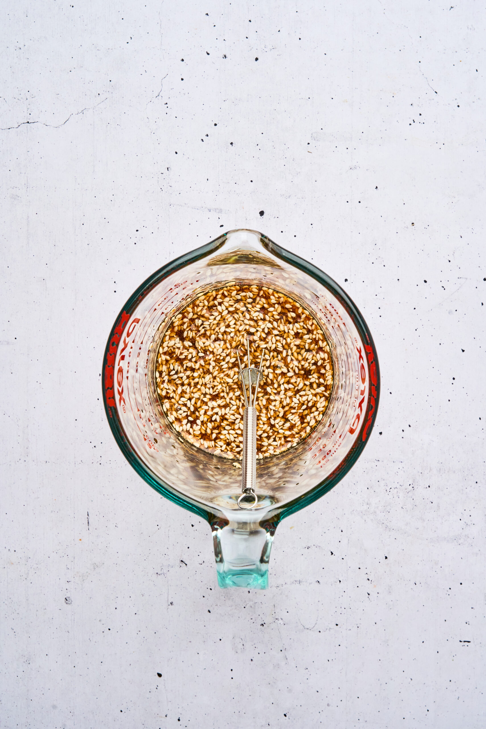 Overhead view of a Pyrex measuring cup filled with sesame seeds and sauce ingredients, with a small whisk inside.