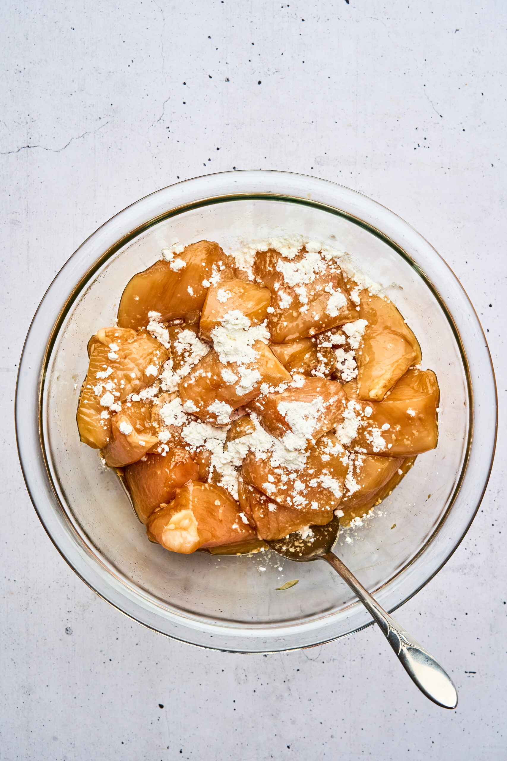 Overhead view of raw chicken pieces in a bowl, topped with cornstarch, with a spoon in the bowl.