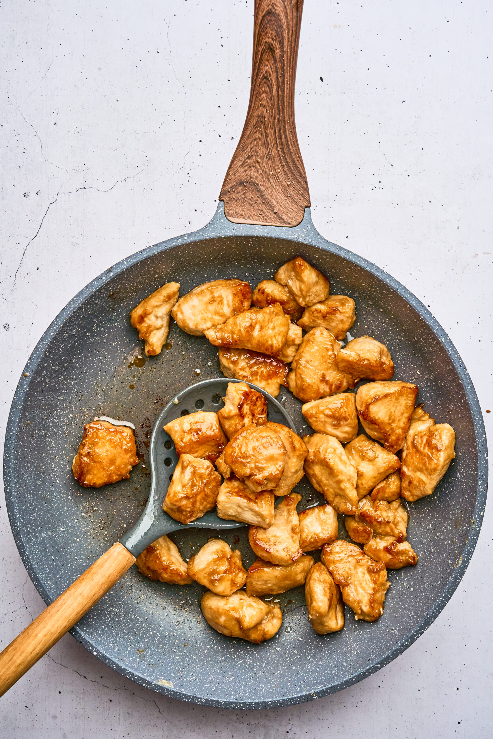 Overhead view of cooked chicken pieces in a frying pan, with a slotted spoon scooping some pieces.