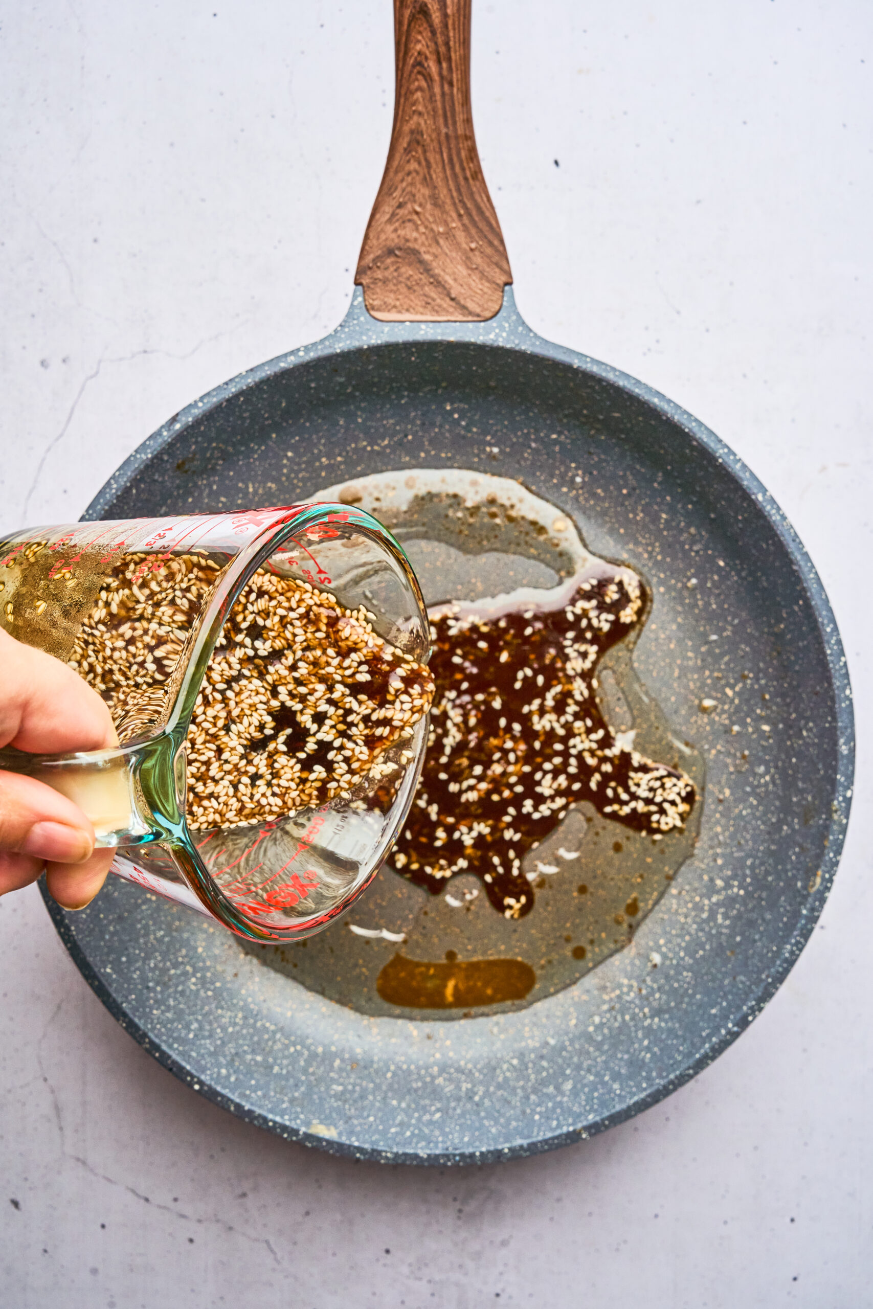 Overhead view of a person pouring a sesame and sauce mixture from a measuring cup into a frying pan.