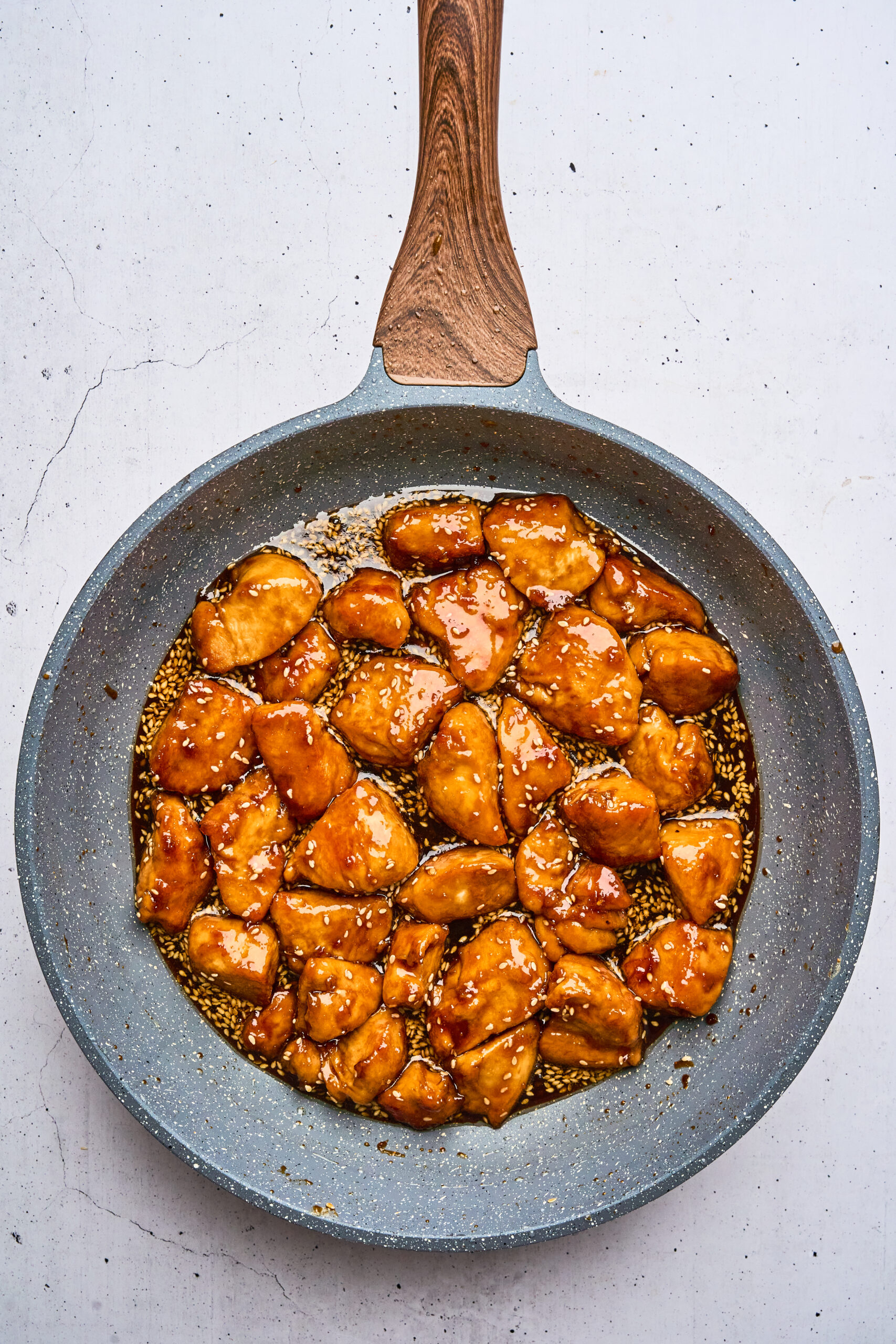 Overhead view of cooked honey sesame chicken in a frying pan, coated with sauce and garnished with sesame seeds.