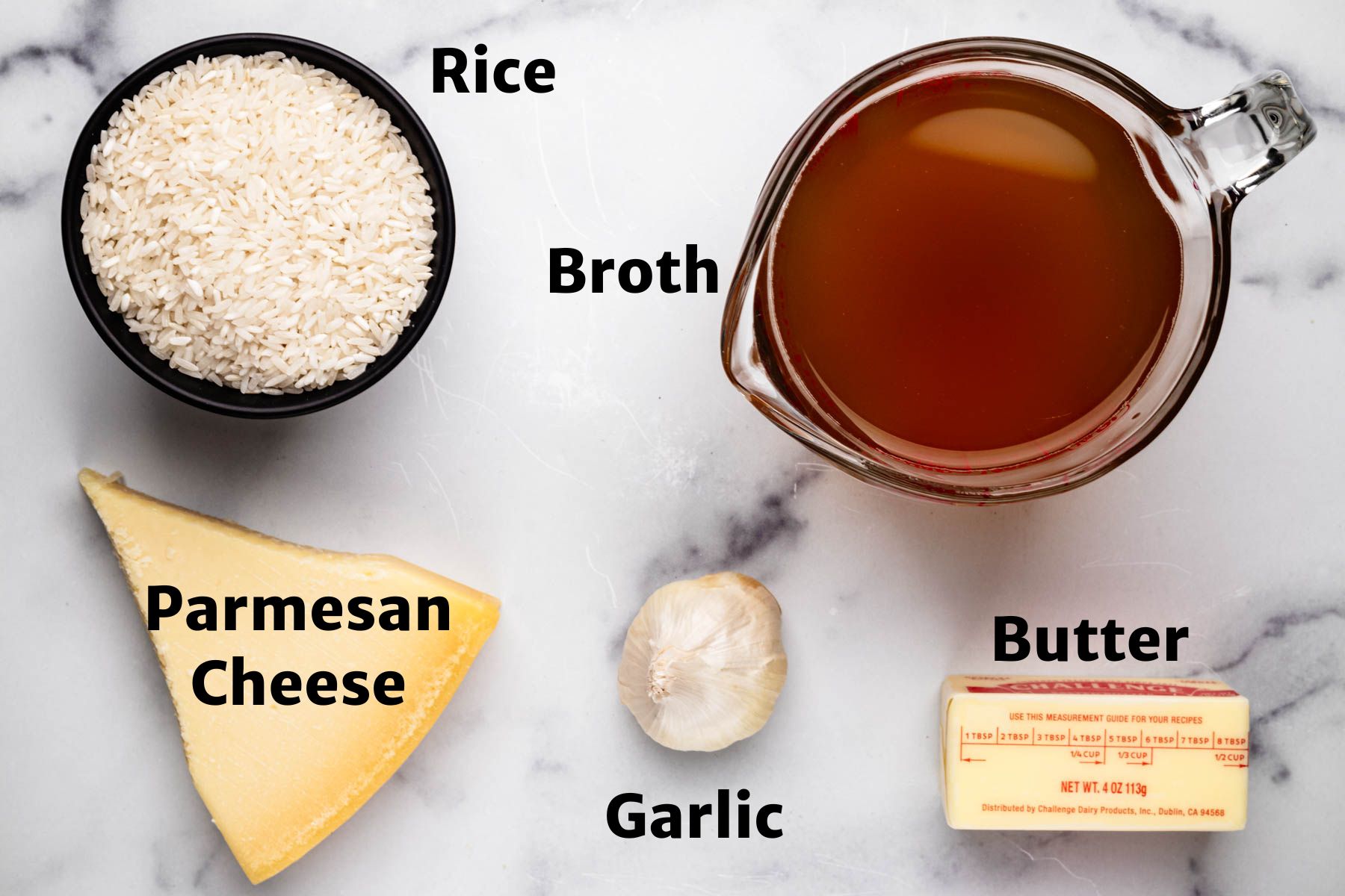 Ingredients for Garlic Parmesan Rice: a black bowl of uncooked rice, a wedge of Parmesan cheese, a garlic bulb, a stick of butter, and a measuring cup of broth arranged on a white marble background.