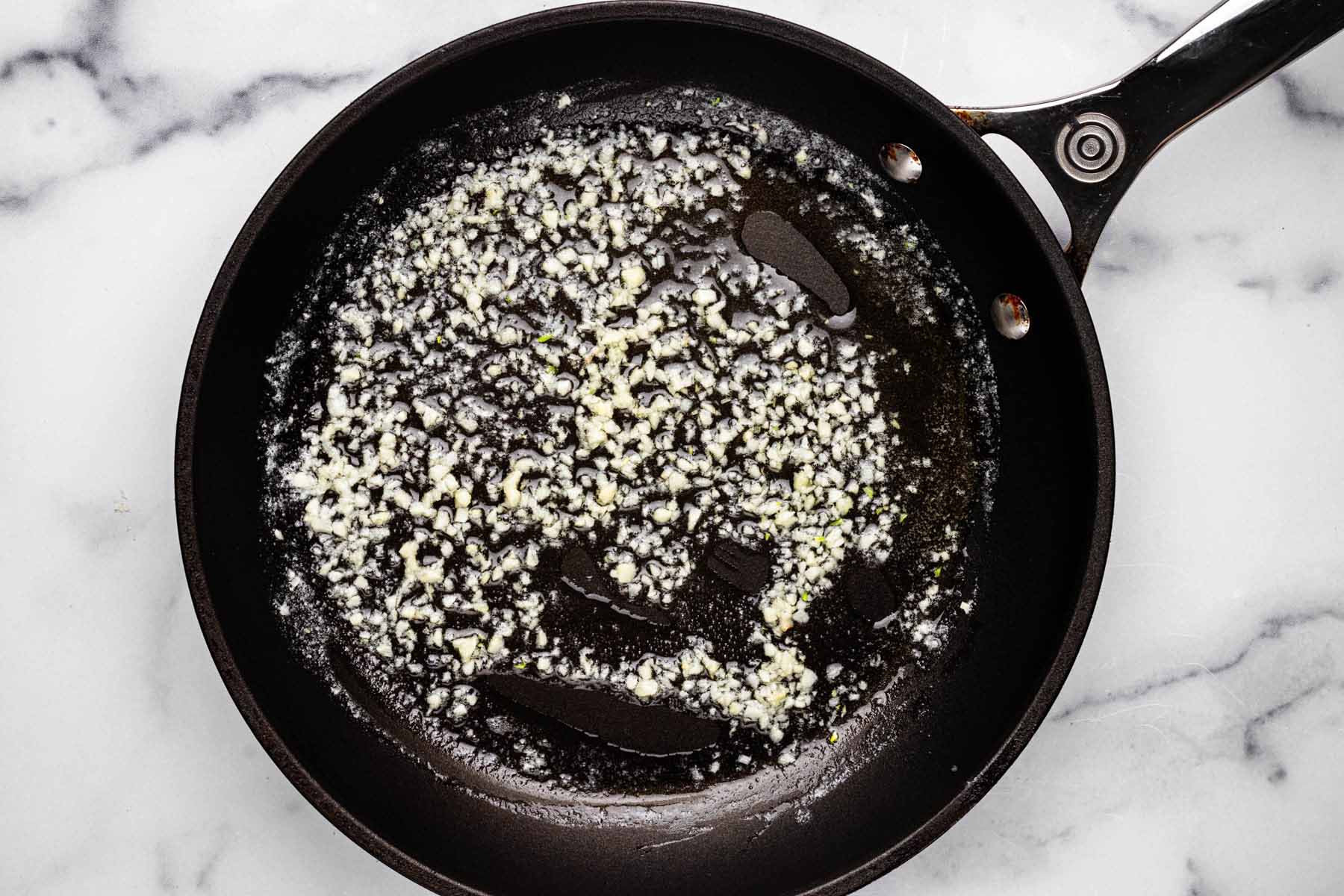 Minced garlic sautéing in melted butter in a black skillet, creating a fragrant base for Garlic Parmesan Rice, set on a white marble background.