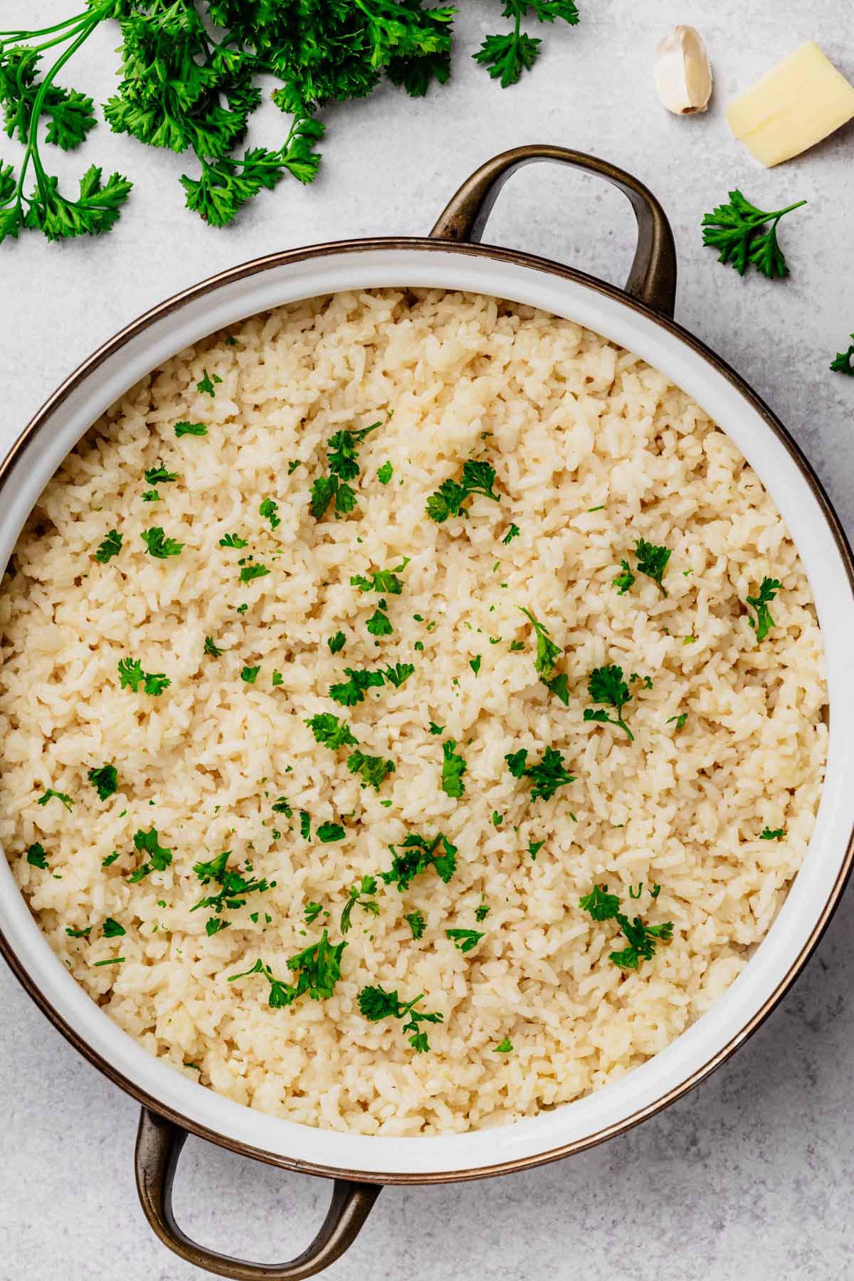 Large serving dish filled with fluffy Garlic Parmesan Rice, garnished with fresh parsley, surrounded by parsley sprigs, a garlic clove, and a pat of butter on a light background.