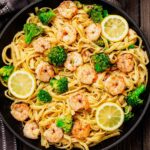 Shrimp and Broccoli Scampi with fettuccine pasta in a black skillet, topped with lemon slices and seasoned broccoli florets, on a rustic wooden background.