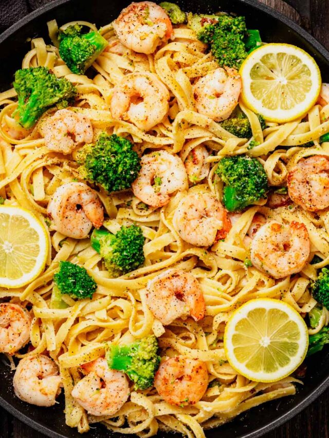 Shrimp and Broccoli Scampi with fettuccine pasta in a black skillet, topped with lemon slices and seasoned broccoli florets, on a rustic wooden background.