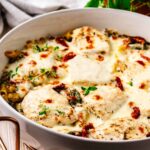 Close-up of a creamy Tuscan chicken bake topped with melted mozzarella cheese, sun-dried tomatoes, and fresh herbs in a white baking dish.