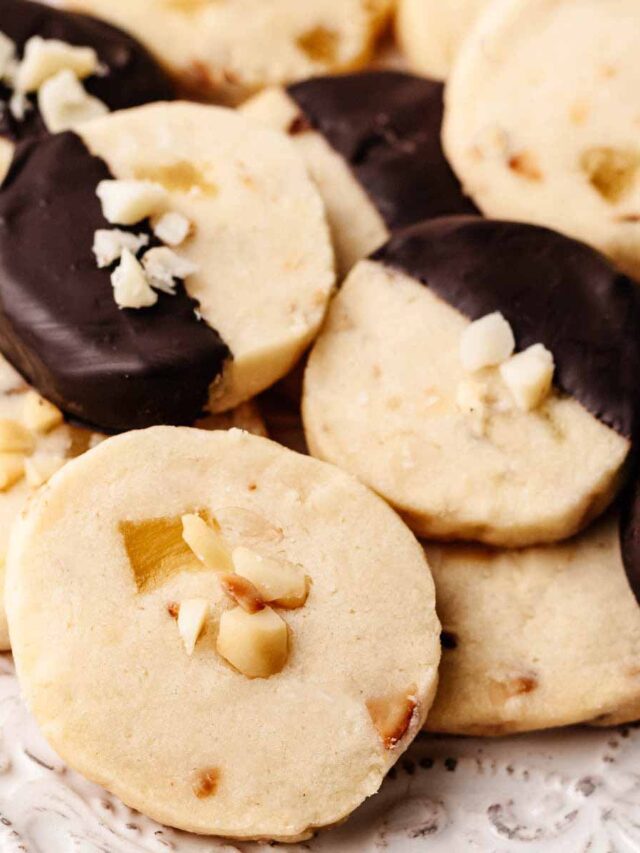 Close-up of Hawaiian shortbread cookies featuring macadamia nuts and dried pineapple, with some cookies partially dipped in dark chocolate and garnished with chopped nuts.