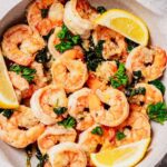 Close-up of lemon butter shrimp in a skillet, garnished with fresh parsley and wilted spinach, with lemon wedges on the side. The shrimp are golden and coated in a buttery sauce.