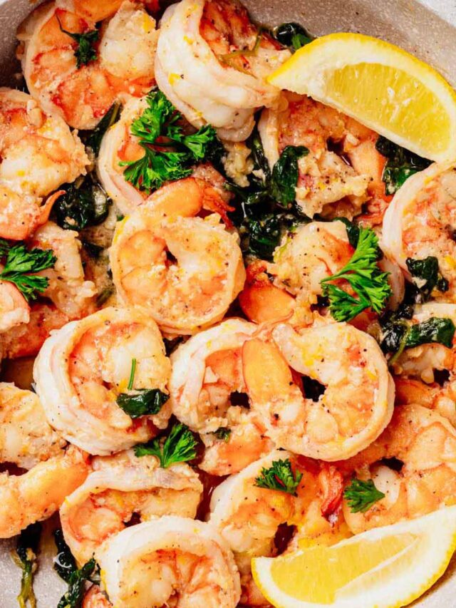 Close-up of lemon butter shrimp in a skillet, garnished with fresh parsley and wilted spinach, with lemon wedges on the side. The shrimp are golden and coated in a buttery sauce.