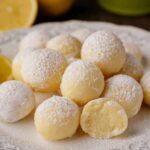 A close-up of Limoncello truffles on a white decorative plate. The truffles are smooth, coated with a light dusting of powdered sugar, and one truffle is cut open to reveal the creamy, lemony interior. Fresh lemons are visible in the background.