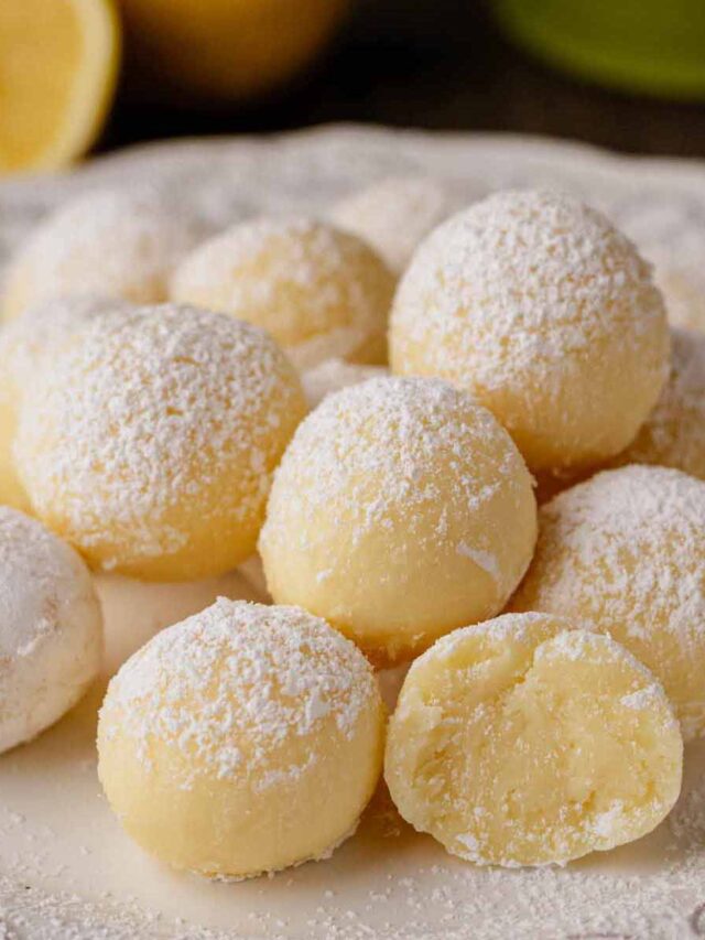 A close-up of Limoncello truffles on a white decorative plate. The truffles are smooth, coated with a light dusting of powdered sugar, and one truffle is cut open to reveal the creamy, lemony interior. Fresh lemons are visible in the background.