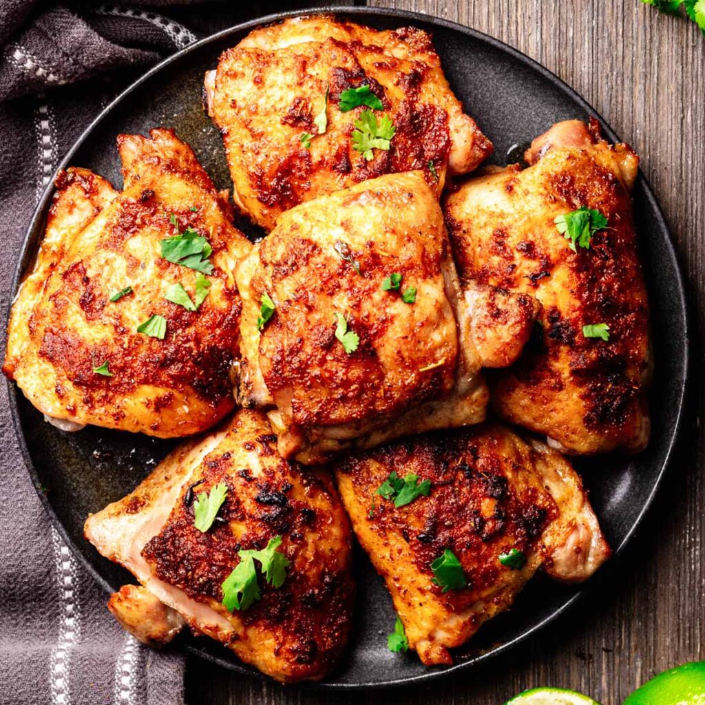 Perfectly baked Mexican chicken thighs with crispy, golden skin, garnished with fresh cilantro and served on a black plate, placed on a rustic wooden surface.