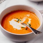 Close-up of creamy roasted red pepper and Gouda soup served in a white bowl, garnished with a swirl of cream and fresh thyme, with a spoon resting inside.