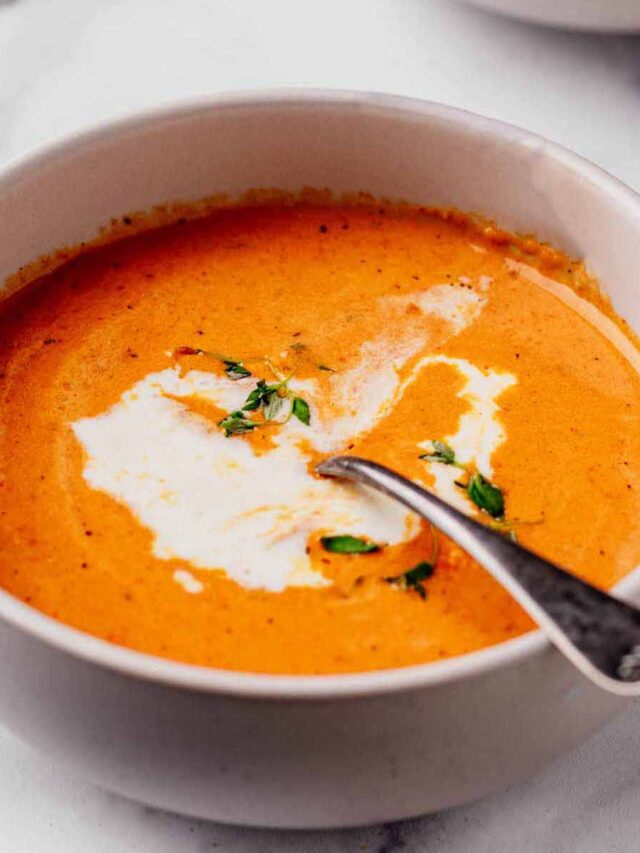 Close-up of creamy roasted red pepper and Gouda soup served in a white bowl, garnished with a swirl of cream and fresh thyme, with a spoon resting inside.