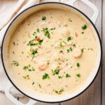 Overhead view of a creamy seafood chowder in a white Dutch oven, featuring shrimp, diced potatoes, and fresh parsley garnish.